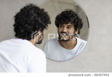 Portrait of happy indian man looking at his reflection in round mirror on the wall, young guy standing in bathroom Portrait of happy indian man looking at his reflection in round mirror on the wall, young guy standing in bathroom 86407491