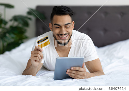 Young Middle Eastern Man Using Digital Tablet And Credit Card In Bed Young Middle Eastern Man Using Digital Tablet And Credit Card In Bed 86407614
