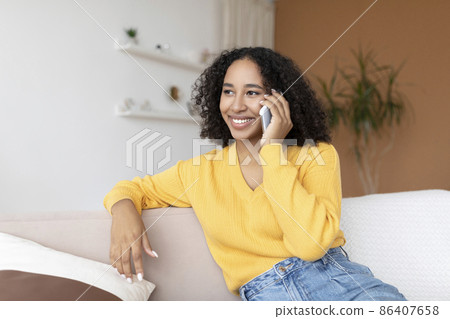 Cheery young African American woman talking on smartphone, sitting on sofa at home Cheery young African American woman talking on smartphone, sitting on sofa at home 86407658