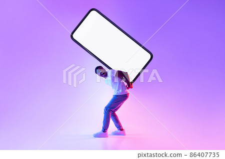 Attractive milennial black guy carrying large heavy smartphone with blank screen on his back in neon light 86407735