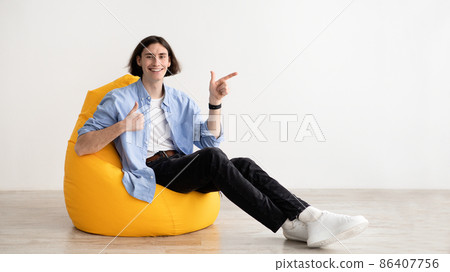 Happy man gesturing thumb up and pointing aside at free space, sitting in beanbag chair on light wall, panorama 86407756