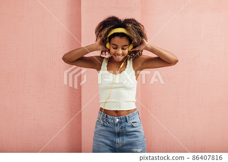 Young smiling woman in casuals with yellow headphones enjoying music while standing indoors at pink wall 86407816