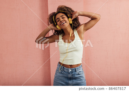 Young smiling woman in casuals with yellow headphones enjoying music while standing indoors at pink wall Young smiling woman in casuals with yellow headphones enjoying music while standing indoors at pink wall 86407817