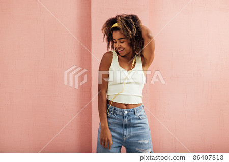 Young smiling woman in casuals with yellow headphones enjoying music while standing indoors at pink wall Young smiling woman in casuals with yellow headphones enjoying music while standing indoors at pink wall 86407818