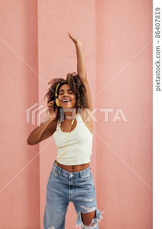 Young smiling woman in casuals with yellow headphones enjoying music while standing indoors at pink wall 86407819
