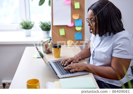 Young black lady is shocked by something during work sitting behind desk with laptop Young black lady is shocked by something during work sitting behind desk with laptop 86409081