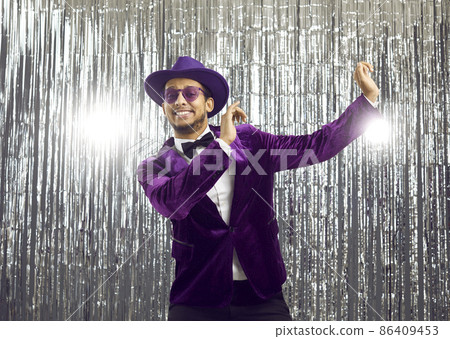 Happy goofy young man wearing a ridiculous tacky unfashionable purple velvet jacket and hat doing funny dance moves at a party. Hilarious dorky guy dancing to music on a silver foil fringe background Happy goofy young man wearing a ridiculous tacky unfashionable purple velvet jacket and hat doing funny dance moves at a party. Hilarious dorky guy dancing to music on a silver foil fringe background 86409453