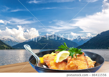 Traditional austrian food (Wiener schnitzel) against Zell am see village with lake in Austria 86411556