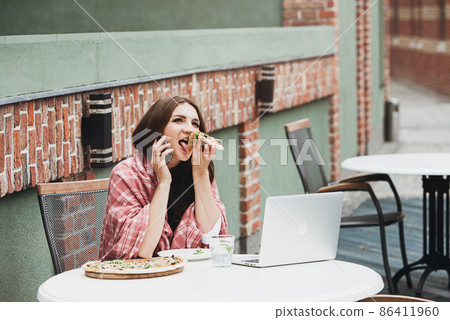 Young freelancer communicates using smartphone while eating pizza in cafe Young freelancer communicates using smartphone while eating pizza in cafe 86411960