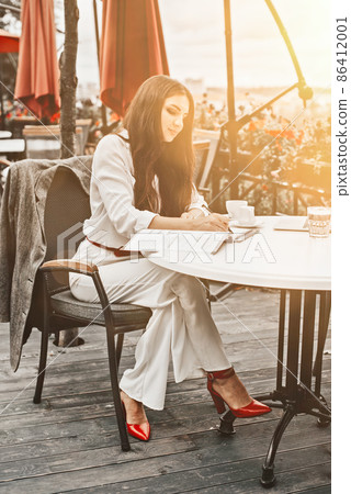 Smiling young dark-haired businesswoman makes notes in a notebook in a cafe on a summer day. Business, e-learning, freelance concept. laptop on a table Smiling young dark-haired businesswoman makes notes in a notebook in a cafe on a summer day. Business, e-learning, freelance concept. laptop on a table 86412001