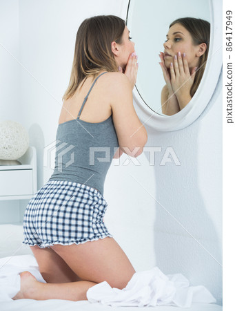 Young female touching face, using mirror in bedroom 86417949