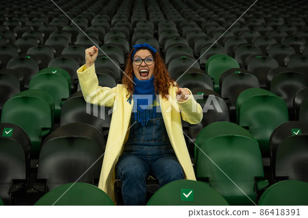 Caucasian woman cheers for a sports team at the stadium. The girl watches the match at the stadium alone. Caucasian woman cheers for a sports team at the stadium. The girl watches the match at the stadium alone. 86418391