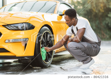 Handsome African guy wiping wheel of modern luxury car with green microfiber cloth Handsome African guy wiping wheel of modern luxury car with green microfiber cloth 86421316