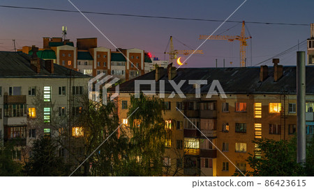 Light in the windows of high-rise buildings at night in summer and half-moon in the sky between houses 86423615