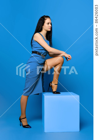 Young woman in blue dress and stockings put her foot on the cube in blue studio Young woman in blue dress and stockings put her foot on the cube in blue studio 86426083