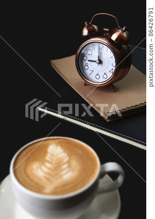 Alarm clock and notepad for notes.The coffee cup is out of focus.Food and drinks.Coffee break. Space for copying.Black background.Vertical photo. Alarm clock and notepad for notes.The coffee cup is out of focus.Food and drinks.Coffee break. Space for copying.Black background.Vertical photo. 86426791