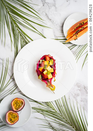 Flat lay of plates with mixed fruits and sliced fruits. Tropical fruit salad served in half dragon fruit. Dragon fruit, maracuja, papaya, palm leaves on table. Top view. Flat lay of plates with mixed fruits and sliced fruits. Tropical fruit salad served in half dragon fruit. Dragon fruit, maracuja, papaya, palm leaves on table. Top view. 86429628