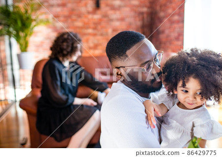 Portrait of upset man hugs his cute curly baby girl in living room Portrait of upset man hugs his cute curly baby girl in living room 86429775