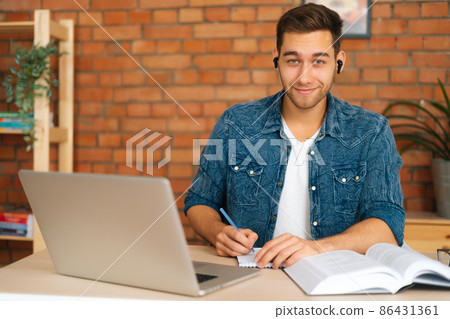 Front view of smiling male student studying online from home using laptop, writing notes, watching video class lesson, listening audio course, studying with teacher distant, lookign at camera. Front view of smiling male student studying online from home using laptop, writing notes, watching video class lesson, listening audio course, studying with teacher distant, lookign at camera. 86431361