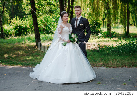 The bride and groom are walking in the park in the summer, hugging. Trees in the background 86432293