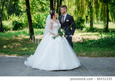 The bride and groom are walking in the park in the summer, hugging. Trees in the background 86432295