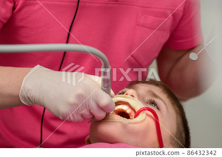 The boy is sitting in a dentist's chair, treating caries in a child, the dentist removes caries from the tooth with a drill. 86432342