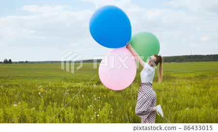 Happy girl with big multicolored balloons posing on the field. 86433015
