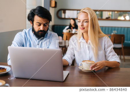 Portrait of two business people of bearded young businessman and pretty Caucasian blonde woman working with laptop in cafe and drinking coffee. Friendly colleagues discussing project in coffee shop 86434840