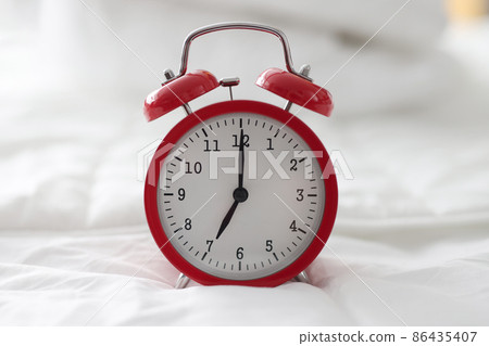 Red alarm clock for 7 hour standing on white bed closeup 86435407