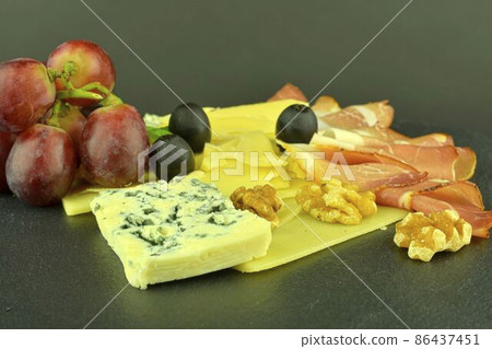 Assorted cheeses on black slate board. Brie cheese, cheddar slices, walnuts, grapes, olives and Schwarzwald ham. Still life food. Copy space Assorted cheeses on black slate board. Brie cheese, cheddar slices, walnuts, grapes, olives and Schwarzwald ham. Still life food. Copy space 86437451