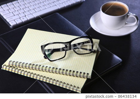 Notepad for notes and glasses are on the table. Black background. The concept of a business breakfast or coffee break. Food and drinks. Computer keyboard. A cup of coffee. Notepad for notes and glasses are on the table. Black background. The concept of a business breakfast or coffee break. Food and drinks. Computer keyboard. A cup of coffee. 86438134