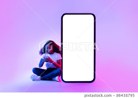 Cheery black woman sitting near giant smartphone with mockup for your app design, using mobile device in neon light 86438795