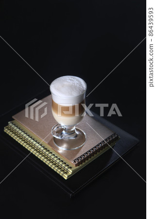 A coffee drink in a tall glass sits on a stack of notebooks. A three-layer drink made of coffee, milk and creamy foam.Concept of a business breakfast or coffee break. Food and drinks. A coffee drink in a tall glass sits on a stack of notebooks. A three-layer drink made of coffee, milk and creamy foam.Concept of a business breakfast or coffee break. Food and drinks. 86439593