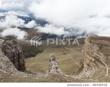 Aerial view of stone amphitheater in thick clouds, Bermamyt plateau, Caucasus. Flying over majestic cliff face along which road runs along the ground. The fog is moving up steep slope. Elbrus region 86440416