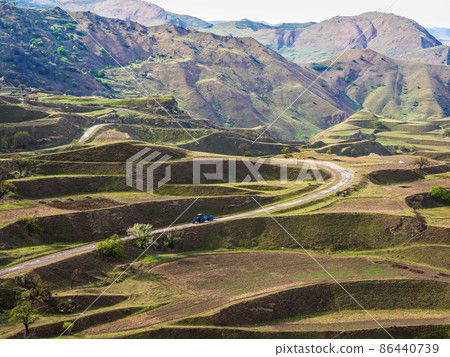 Terraces of spring mountain agriculture. Old tractor is driving along a winding mountain dirt road. Dangerous narrow mountain road through the hills to a high-altitude village. Terraces of spring mountain agriculture. Old tractor is driving along a winding mountain dirt road. Dangerous narrow mountain road through the hills to a high-altitude village. 86440739
