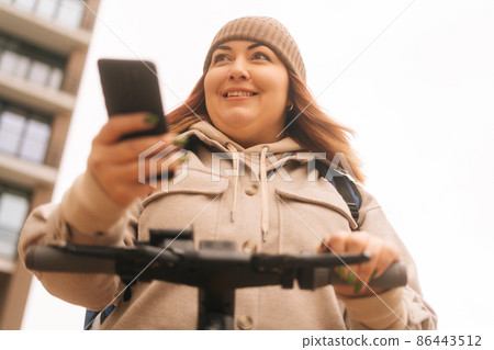 Close-up low-angle view of smiling woman wearing warm hat and jacket using mobile phone standing with electric scooter at urban street in cloudy autumn day, selective focus, blurred background. 86443512