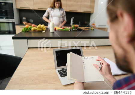 Soft focus on a notepad in the hands of a man working on laptop on the background of his out of focus pregnant wife preparing healthy meal on the kitchen island. Domestic life concept 86444155