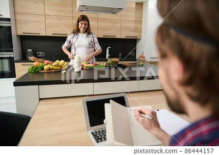 Beautiful pregnant woman smiles toothy smile while chopping vegetables on a cutting board and looking at her blurred husband working from home on laptop and holding a notepad in his hands 86444157