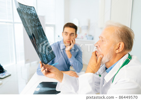 Thoughtful mature adult male physician consult frustrated young man patient giving bad news explaining results of MRI image. Unhappy scared young man listening to bad news sitting in doctor office. 86449570