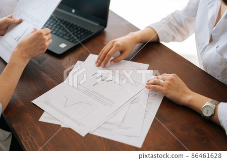 Close-up high-angle view of hands unrecognizable two young business women sitting at desk with laptop and paper documents, discuss project, brainstorming working together. 86461628