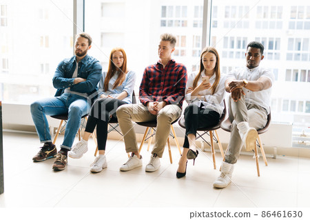 Group of bored young diverse multiracial job candidates in casual clothes waiting interview with hr, sitting in queue line row on chairs in modern office lobby on background of window and cityscape 86461630