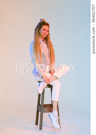 Young woman with dreadlocks in jeans and jacket sits on chair under colorful lights Young woman with dreadlocks in jeans and jacket sits on chair under colorful lights 86462707