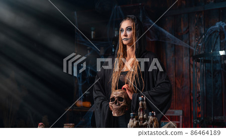 Halloween concept. Witch portrait close up with dreadlocks looking away dressed black hood standing dark room with cage on background 86464189
