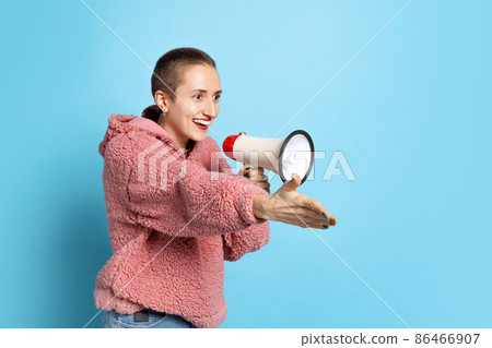 Excited young girl, student with short hair wearing pink hoodie shouting at megaphone isolated on blue background. Concept of emotions, music Excited young girl, student with short hair wearing pink hoodie shouting at megaphone isolated on blue background. Concept of emotions, music 86466907