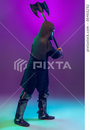 Studio shot of young bearded man in image of medieval warrior, archery in chain armor with ax isolated over purple background in neon. Comparison of eras 86468292