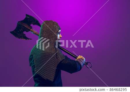Studio shot of young bearded man in image of medieval warrior, archery in chain armor with ax isolated over purple background in neon. Comparison of eras Studio shot of young bearded man in image of medieval warrior, archery in chain armor with ax isolated over purple background in neon. Comparison of eras 86468295