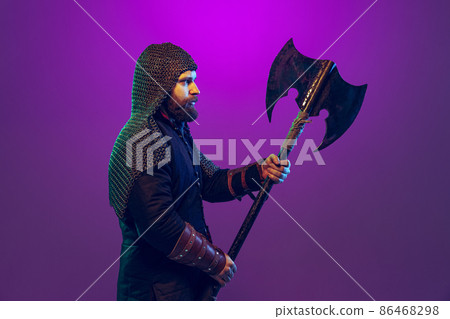 Studio shot of young bearded man in image of medieval warrior, archery in chain armor with ax isolated over purple background in neon. Comparison of eras 86468298