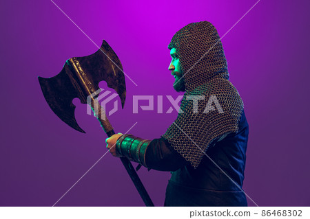 Studio shot of young bearded man in image of medieval warrior, archery in chain armor with ax isolated over purple background in neon. Comparison of eras Studio shot of young bearded man in image of medieval warrior, archery in chain armor with ax isolated over purple background in neon. Comparison of eras 86468302