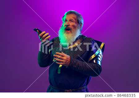 Comic portrait of senior grey-bearded man in image of medieval warrior in armor using smartphone isolated over purple background in neon. Comparison of eras 86468335