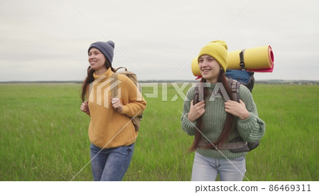 Girl friends with backpacks walk across field hiking Girl friends with backpacks walk across field hiking 86469311